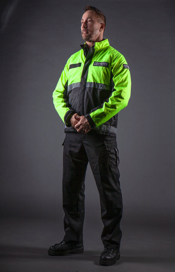 Male model wearing a standard length, waterproof, long sleeve jacket over tactical pants, from a left front angle, with upright collar and no rain-cap.Bottom half is black, topped by fluorescent green top, shoulders, and top of arms. Underarms, wristbands, front zipper, inner lining in collar, velcro patch on both chests and left shoulder are black. Two 2” reflective horizontal stripes cross the jacked mid and bottom. Logo patch on both chests and left shoulder.