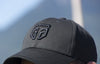 Black cotton baseball cap with a black GTG embroidered logo on barely showing human head, seen from front-left angle. 