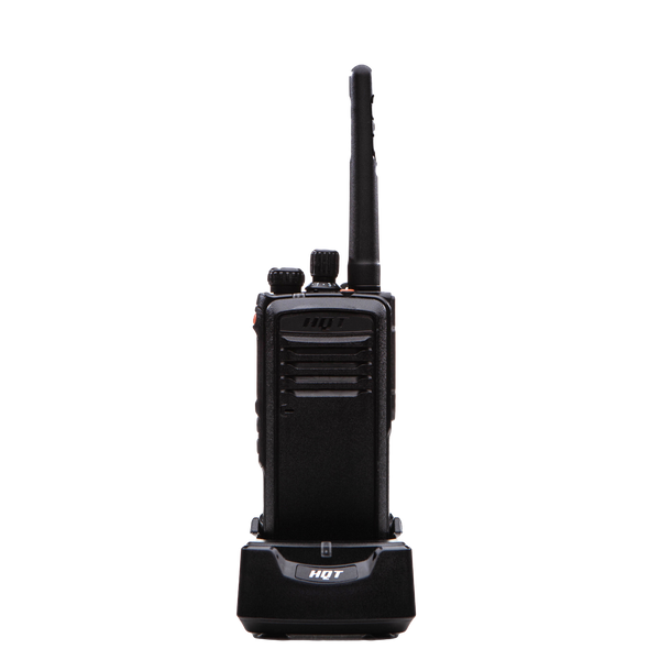 Picture of the front of a black radio, branded with silver HQT on top, resting in its black charger displaying the same silver brand.  On top of the radio one can see a left on/off-volume button, the front LED light, a channel selection button, a red front top key and a short antenna. 