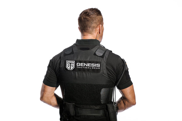 Picture of the back of a black stab-proof vest over a black golf shirt on a human model. A large black embroidered patch displays “GTG GENESIS TACTICAL GEAR” in white on the back. The flaps come from the front to attach on Velcro patches at the back, over the shoulders and at arms height, to attach on the Velcro in the middle back. A belt with accessories pockets is worn over the vest.  An ear piece is worn at the back of the right ear and a remote mic coiled cable appears over the right upper arm.