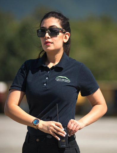 Female model in sunglasses wearing the black short-sleeve golf shirt with three buttons. The left chest has an embroidered logo displaying GENESIS in white, topped by a fluorescent green moon crescent.