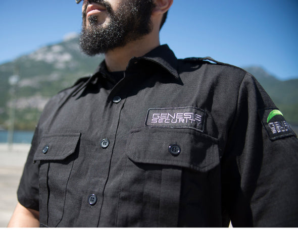 Picture of the left-front of a black tactical shirt with an unbuttoned collar, two buttoned front folded pockets and two shoulder straps, worn by a bearded human model, cut below the chest and at the middle of the head. One embroidered shoulder patch displays GENESIS in white, with a fluorescent green moon crescent atop, and one chest patch displaying only the white GENESIS name.