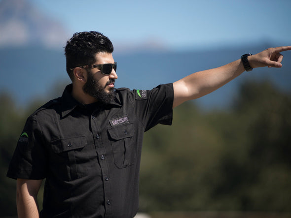 Picture of the front-right angle of a black tactical shirt with an unbuttoned collar, two buttoned front folded pockets and two shoulder straps, worn by a bearded human model wearing sunglasses, pointing to the right with his left hand, cut at the waist. Two embroidered shoulder patches display GENESIS in white, with a fluorescent green moon crescent atop, and one chest patch displays only the white GENESIS name.
